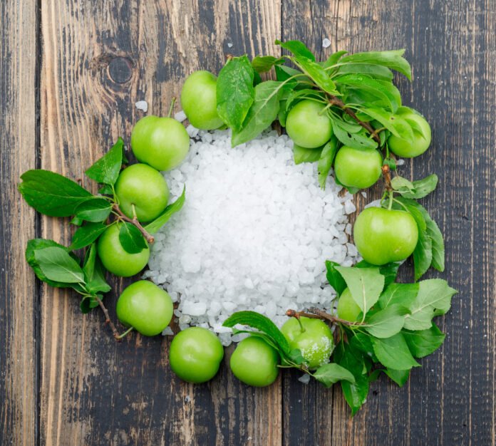 Green plums with salt crystals, leaves on wooden background, flat lay.