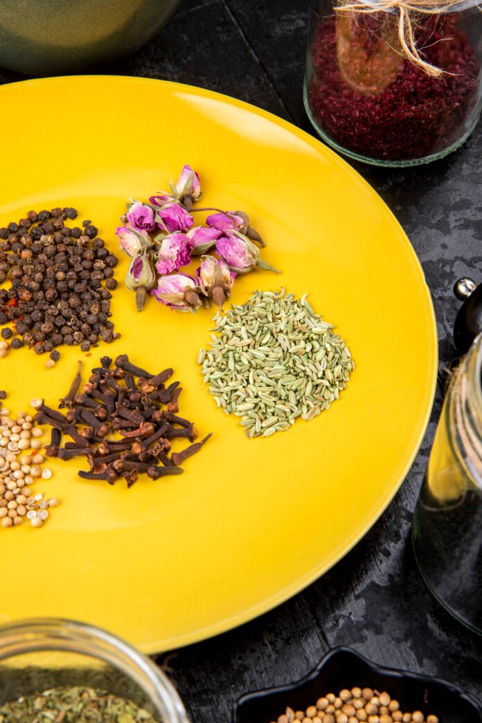 side view of a set of spices and herbs peppercorns anise seeds clove and tea rose buds on yellow plate