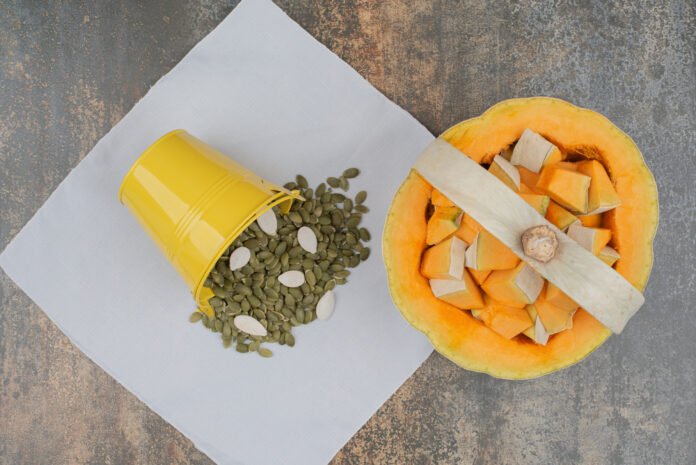 Yellow bucket full of pumpkin peeled seeds with fresh sliced pumpkin on marble background