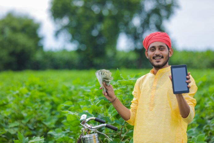 Young indian farmer showing smartphone and money at cotton field